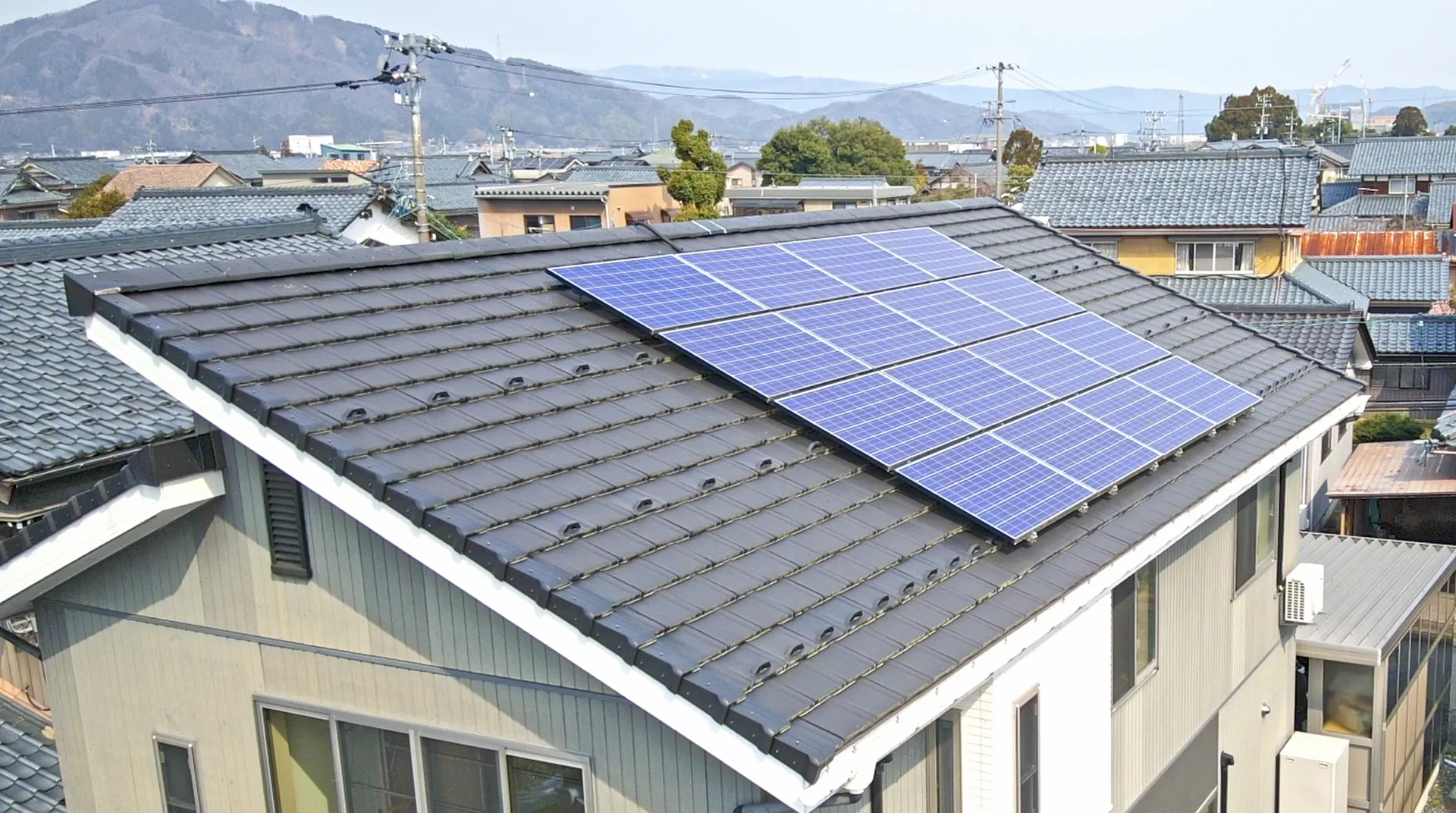 瓦屋根に太陽光パネルが並ぶ2階建て住宅を上から見た様子。周りは住宅街で遠くに山