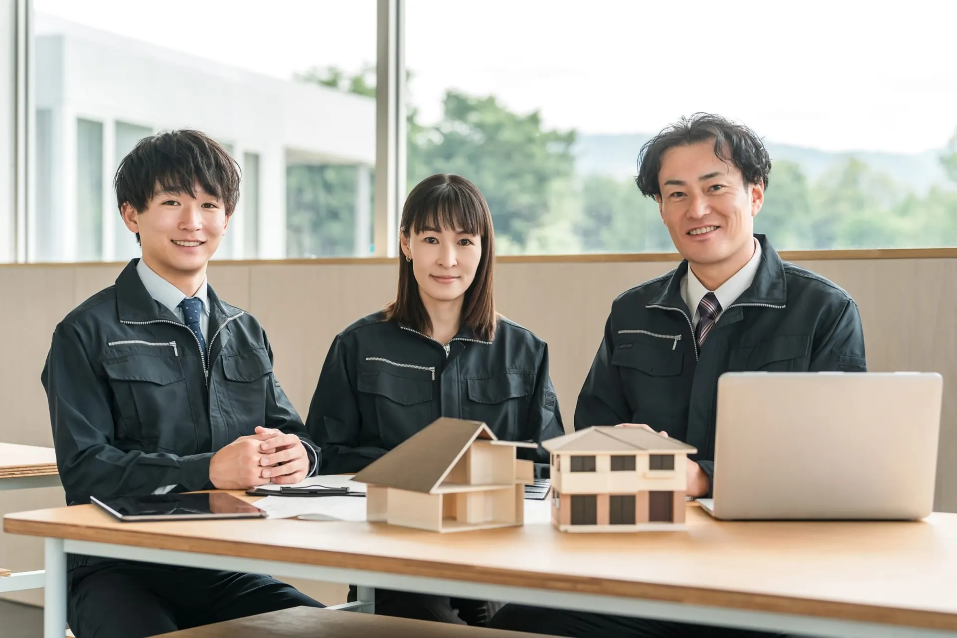 打ち合わせスペースに並ぶ住宅会社のスタッフ3名と、テーブルに置かれた2棟の住宅模型・図面・ノートパソコン