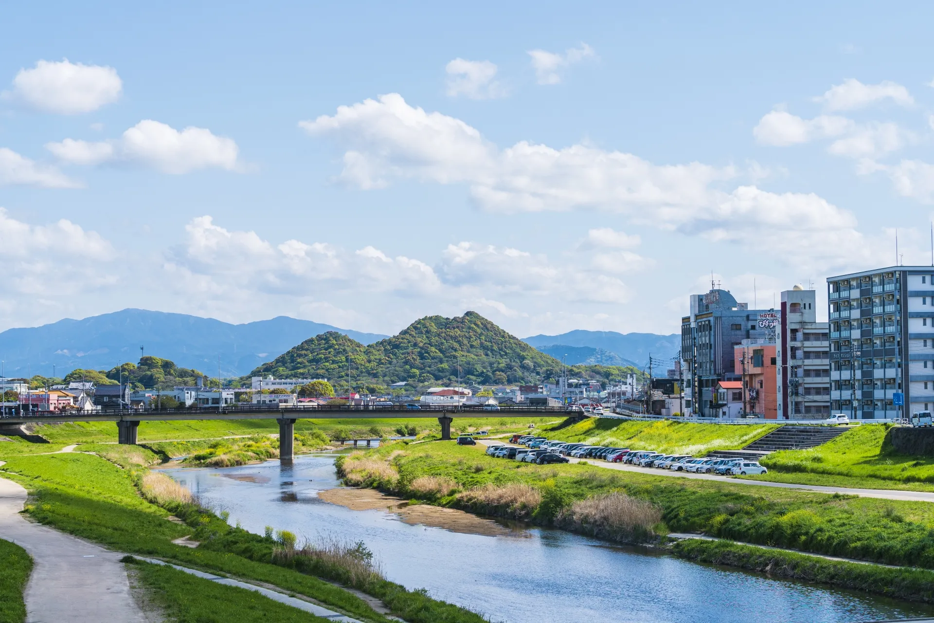 飯塚市の川沿いの風景。堤防の緑地と橋、右手にビルやマンションが並び、背景に低い山並みが見える