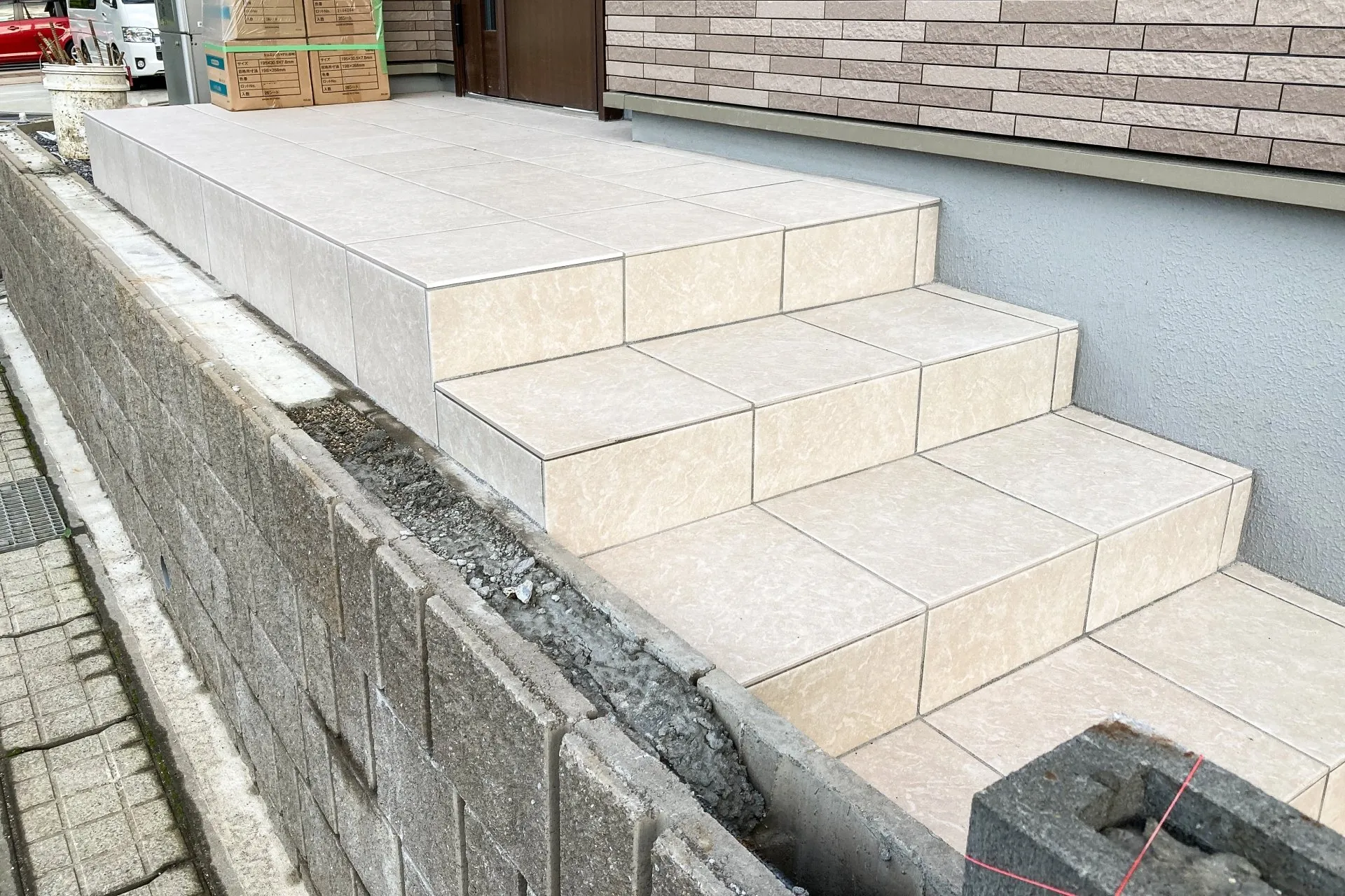 新築住宅の外構工事が進行中の玄関アプローチ。タイル張りの階段が仕上がり、脇の目地はまだ施工途中で、タイルの資材箱が積まれている