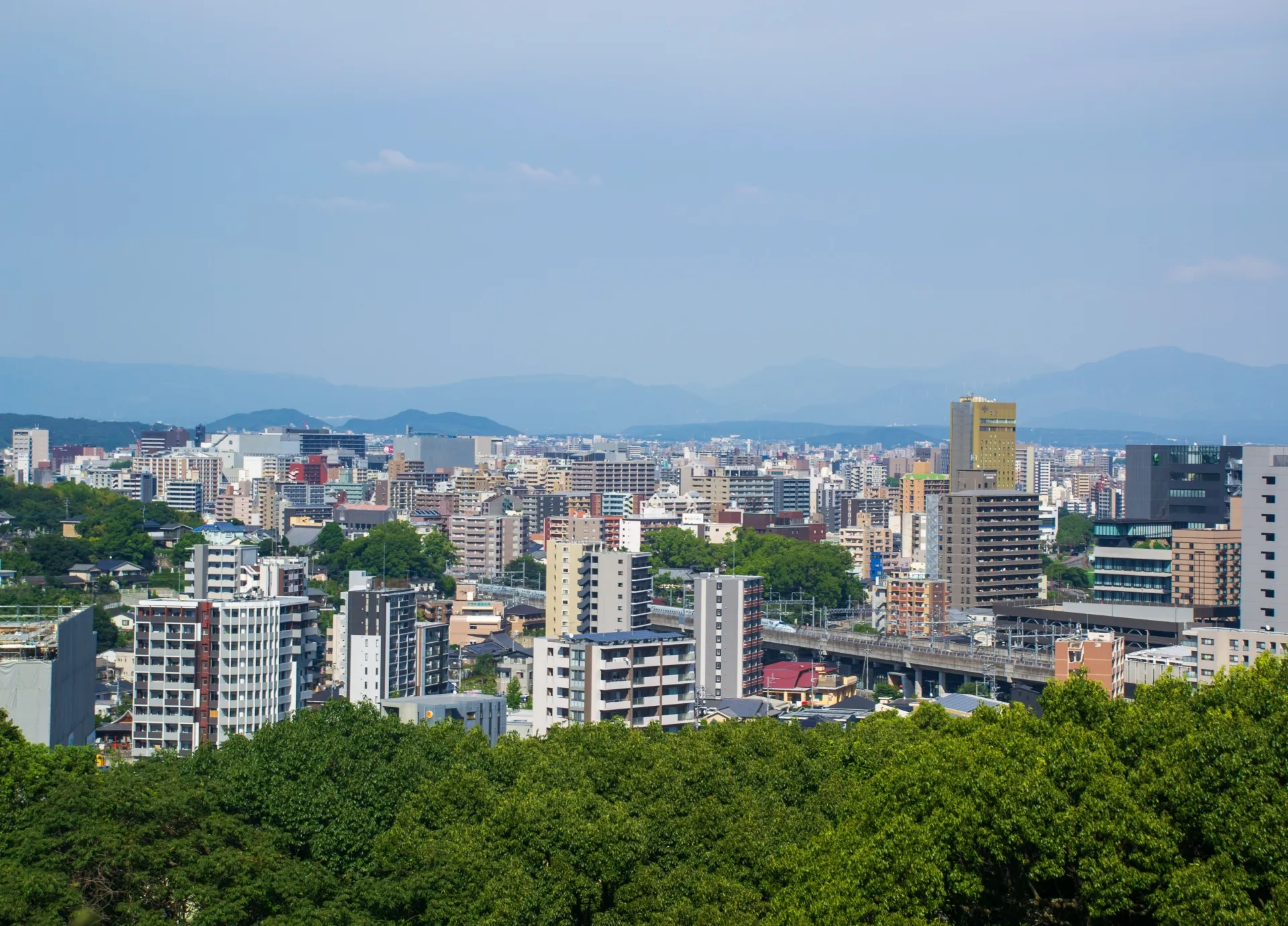 高台から望む熊本市の市街地。中高層のマンションやビルが並び、手前に緑の木々、中央を高架の線路が横切り、遠くに山並みが連なる