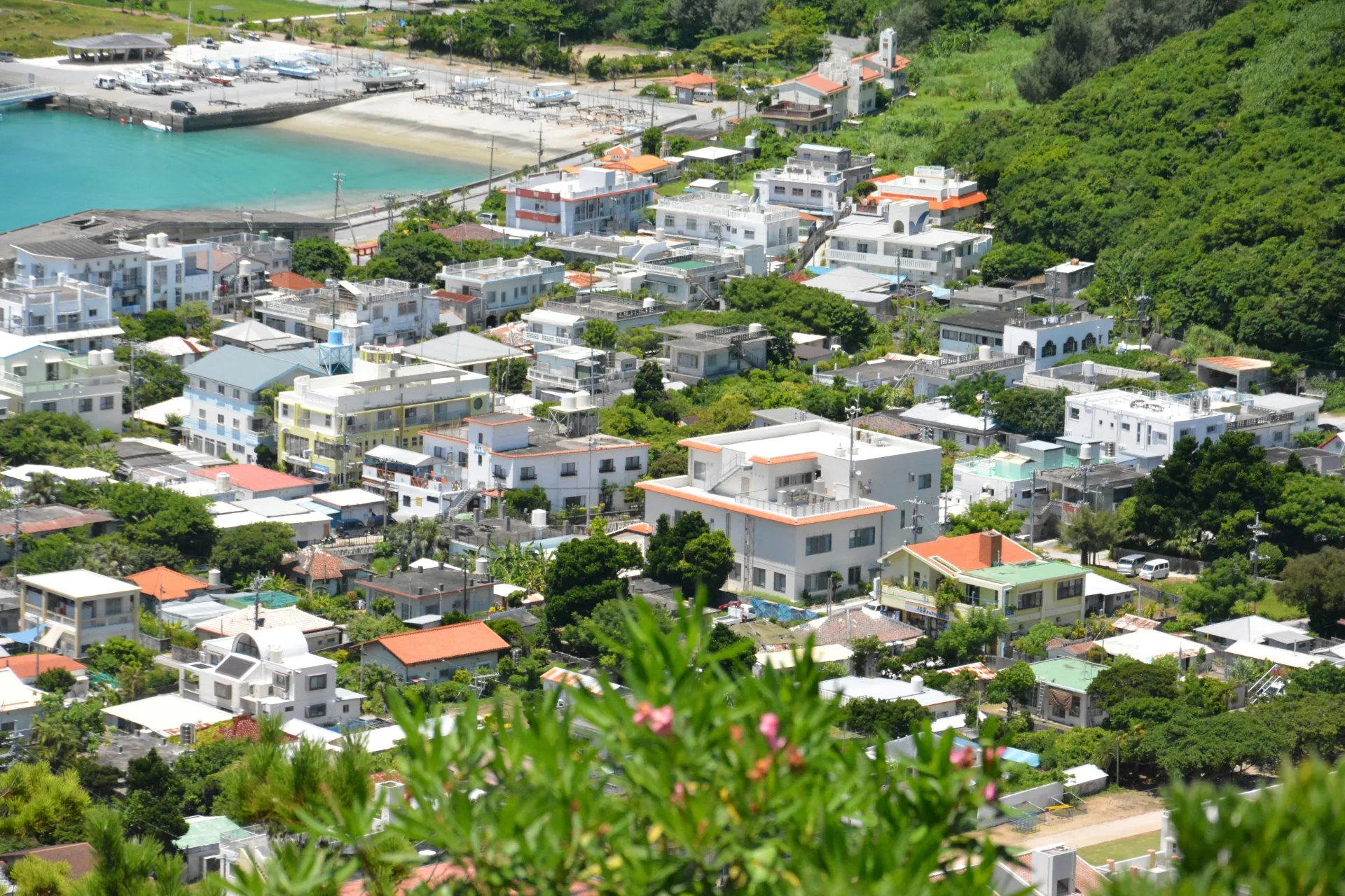 高台から見た沖縄の住宅地。白い外壁と陸屋根の家が並び、左奥に船が並ぶ港と海が見える